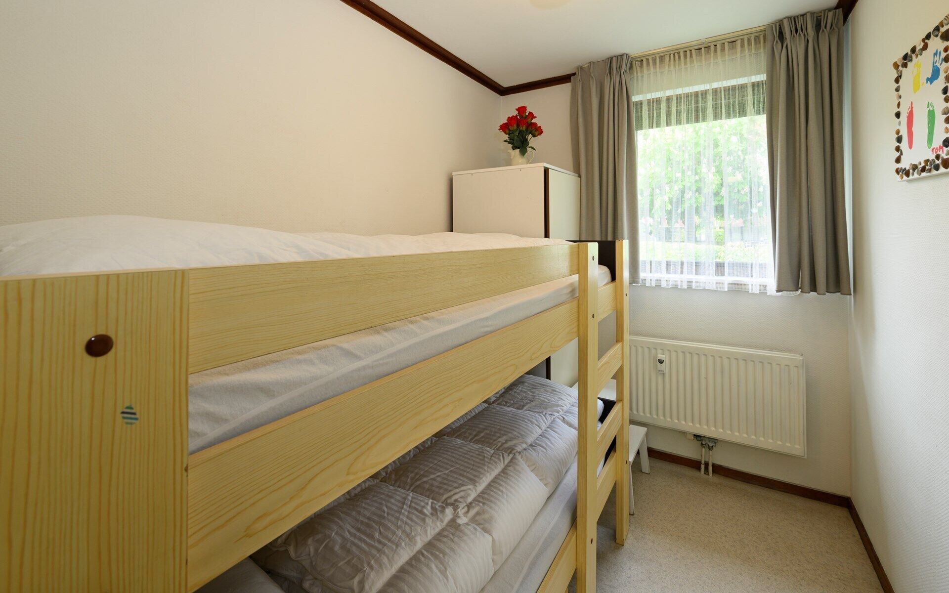 Ein Etagenbett in einem Raum mit Heizkörper, einem Vorhangfenster und einem kleinen Tisch mit Blumen.