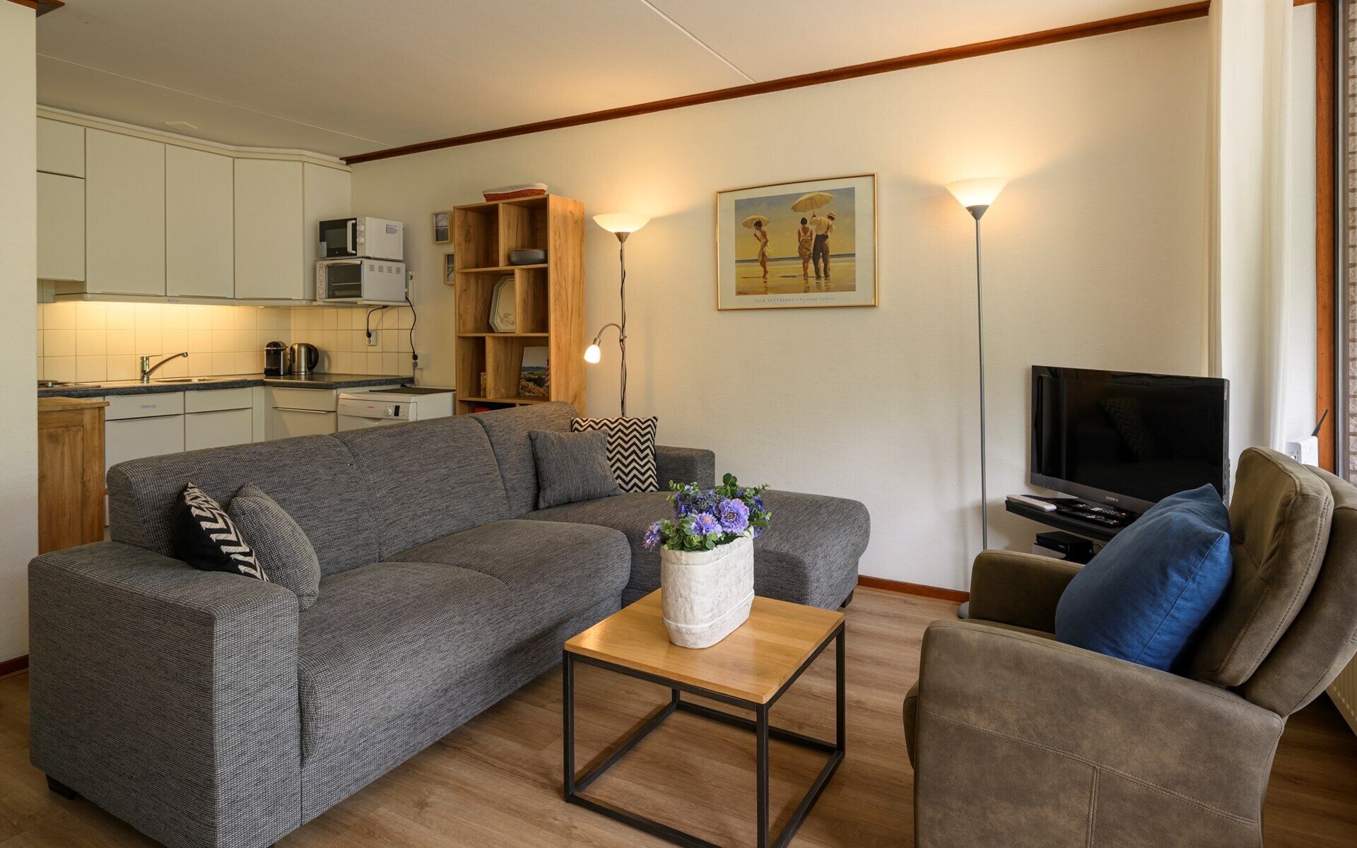 Een woonkamer met een grijze bank, een houten salontafel, een televisie en een keuken op de achtergrond.