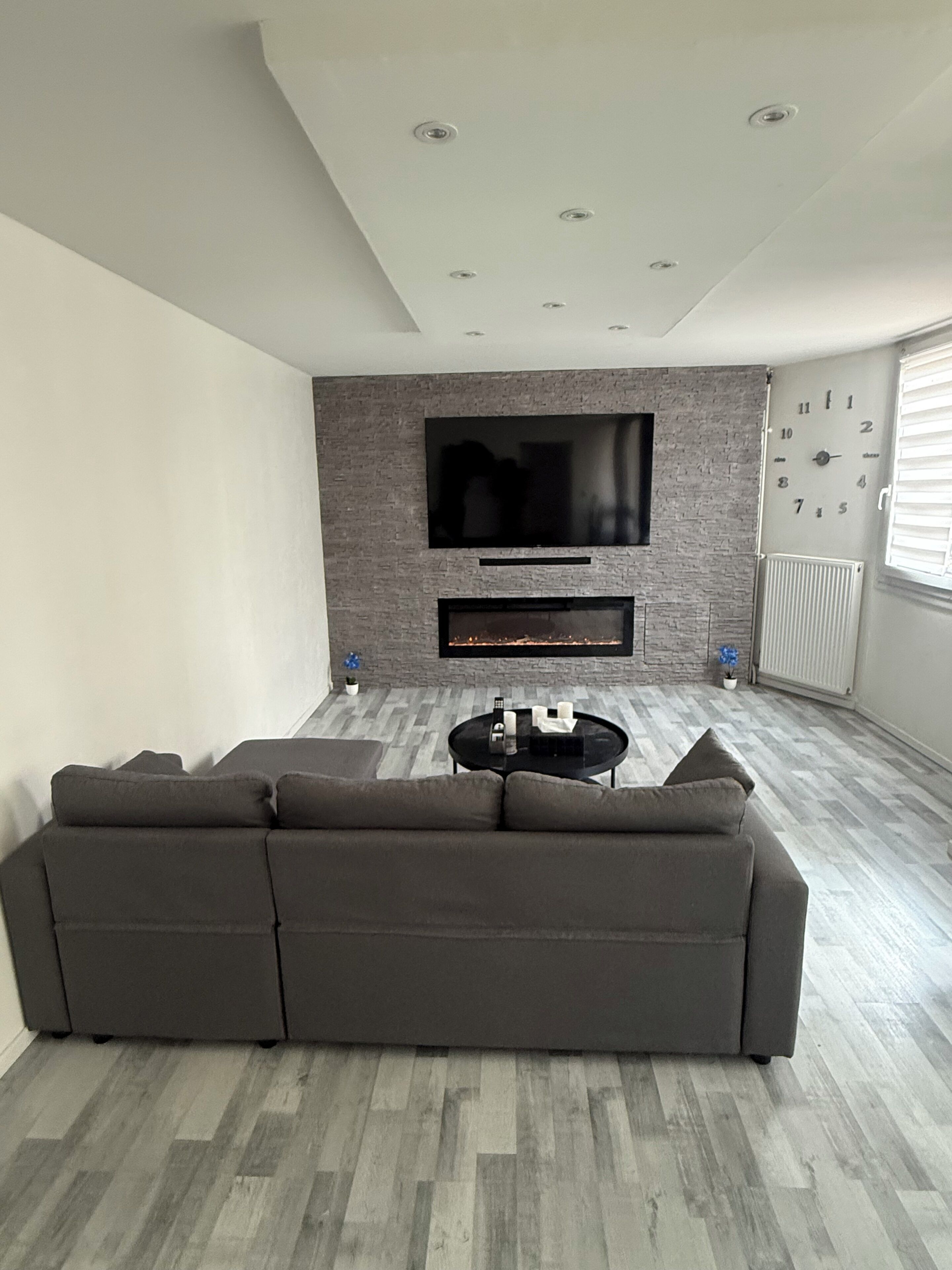 A modern living room with a sectional sofa, a flat-screen TV mounted on a brick wall, a fireplace below the TV, and a radiator on the wall.