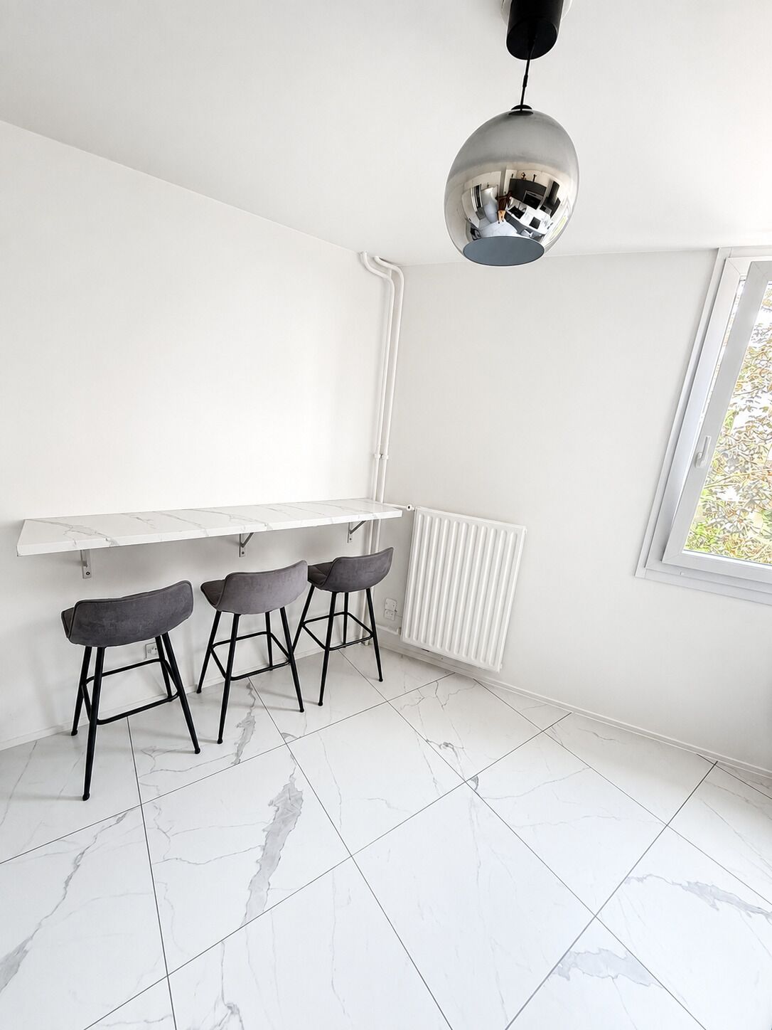 Een moderne keuken met een wit aanrecht, drie barkrukken, een hanglamp en een raam met uitzicht.