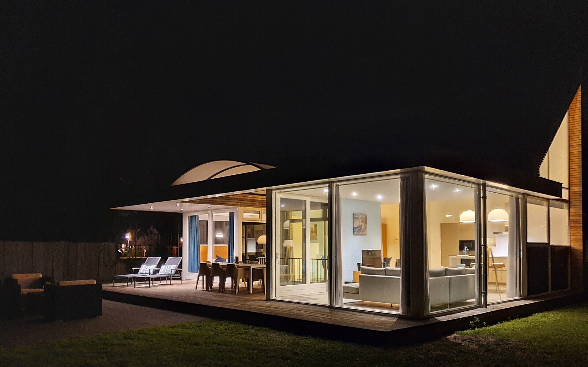 A modern glass house with a deck, outdoor seating, and a visible interior with a sofa and table.
