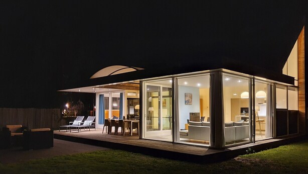 Ein modernes Haus aus Glas mit einer Terrasse, Sitzgelegenheiten im Freien und einem einsehbaren Innenraum mit Sofa und Tisch.