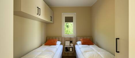 A small, narrow room with two single beds, a window with a view of greenery, and a ceiling light.