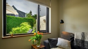 Una habitación con un ventanal grande, una silla, una pequeña mesa con un florero, una lámpara y un sofá con una manta.
