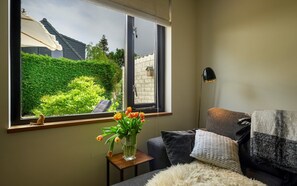 Ein Zimmer mit großem Fenster, einem Sessel, einem kleinen Tisch mit Blumenvase, einer Lampe und einem Sofa mit Überwurfdecke.
