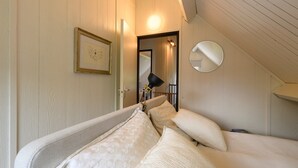 Un dormitorio con una cama, almohadas, una lámpara de noche, un cuadro enmarcado, un espejo y una puerta que da a otra habitación.