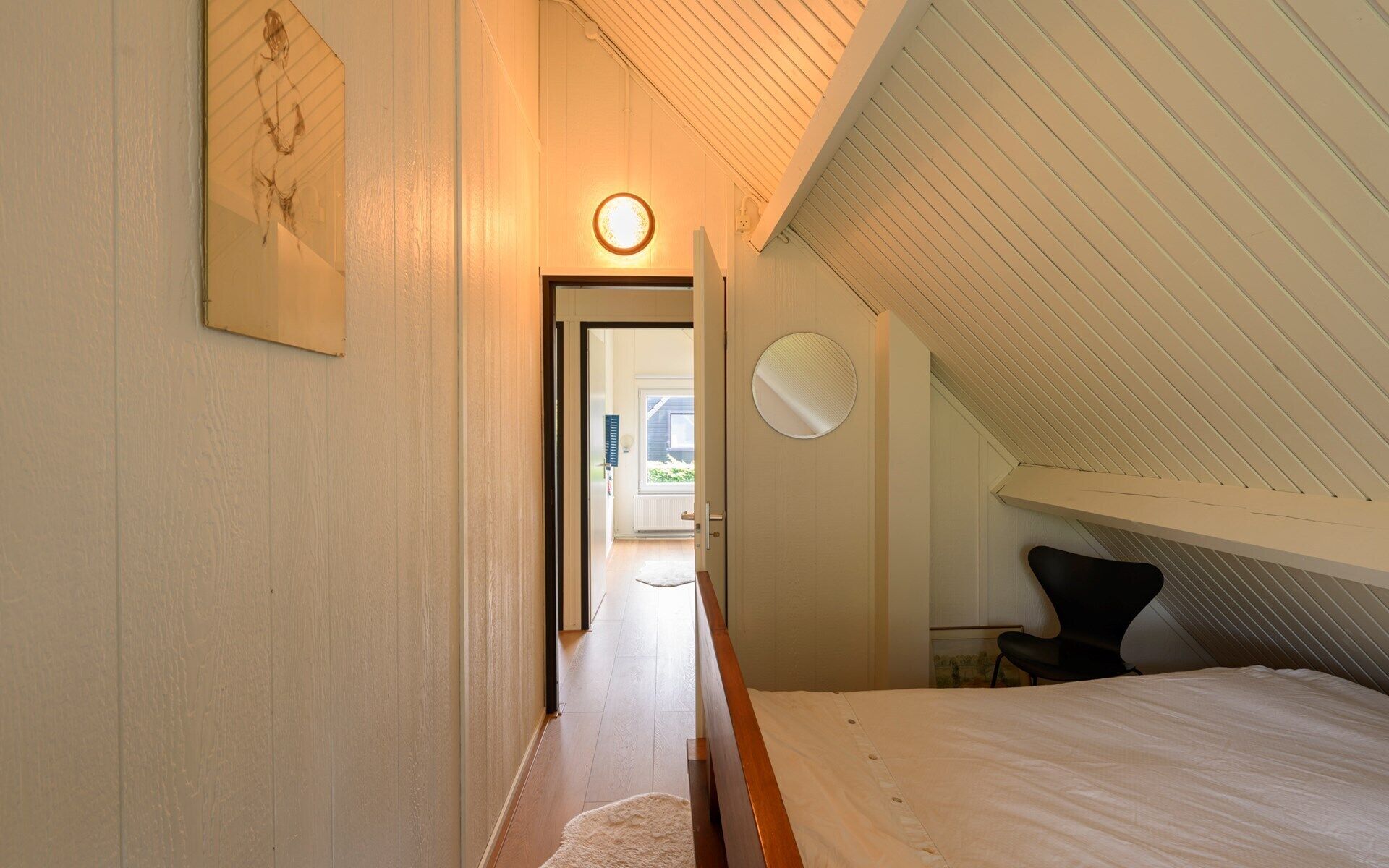 Une chambre avec un plafond en pente, un lit, une chaise et une porte donnant sur un couloir.