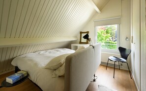 Ein Schlafzimmer mit schrägem Dach, einem Bett, einem Sessel, einem Nachttisch mit Lampe und einem Fenster mit Blick auf Grünflächen.