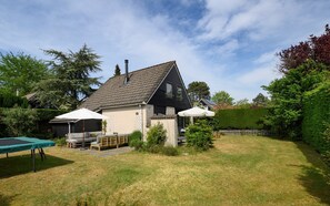 Ein Haus mit geneigtem Dach, einem Schornstein, einem kleinen Garten mit Trampolin und einem weißen Zaun.