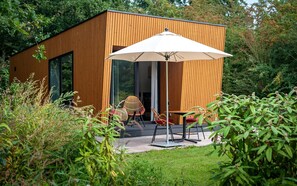 A modern wooden building with a glass door, an outdoor seating area with chairs and a table, and a large umbrella providing shade. - Hoeve Lodge "De Meidoorn" (Burgh-Haamstede)