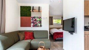 A modern living room with a sectional sofa, a flat-screen TV, a decorative wall panel, and a view into the bedroom. - Hoeve Lodge "De Meidoorn" (Burgh-Haamstede)