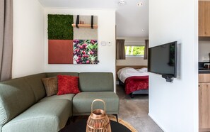 A modern living room with a sectional sofa, a flat-screen TV, a decorative wall panel, and a view into the bedroom. - Hoeve Lodge "De Meidoorn" (Burgh-Haamstede)