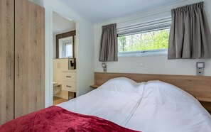Un dormitori amb un llit, una finestra amb cortines, un armari de fusta i un bany visible a través d'una porta oberta.