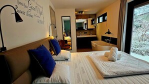 A hotel room with a bed, a nightstand, a lamp, a view of the outside, and a bathroom with a bathtub.