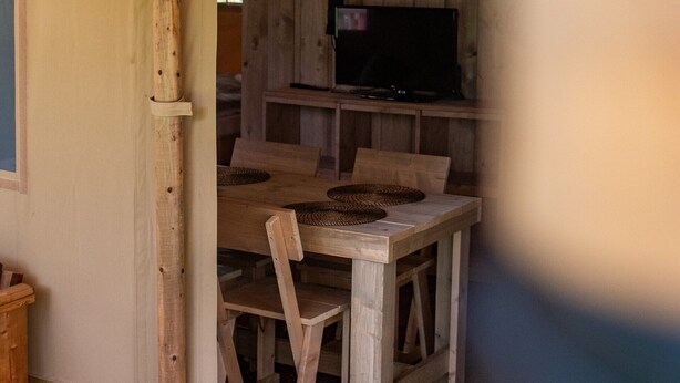 A wooden dining table with woven placemats and chairs, a television on a stand, and a wooden cabinet in the background. - Luxury Safari Tent Renesse (Renesse)