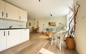 A modern kitchen with white cabinets and a wooden floor, leading to a living area with a sofa and a dining table. - Cottage in the Orchard (Burgh-Haamstede)