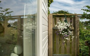 A wooden gate with a hanging flower basket, a window with a view of greenery, and a clear sky. - Cottage in the Orchard (Burgh-Haamstede)
