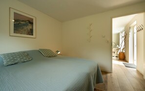 A bedroom with a bed, pillows, a nightstand, a lamp, a painting, and a door leading to another room. - Cottage in the Orchard (Burgh-Haamstede)