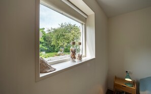 A room with a window overlooking a garden, a wooden nightstand with a lamp, and two glass bottles with cork stoppers on the windowsill.