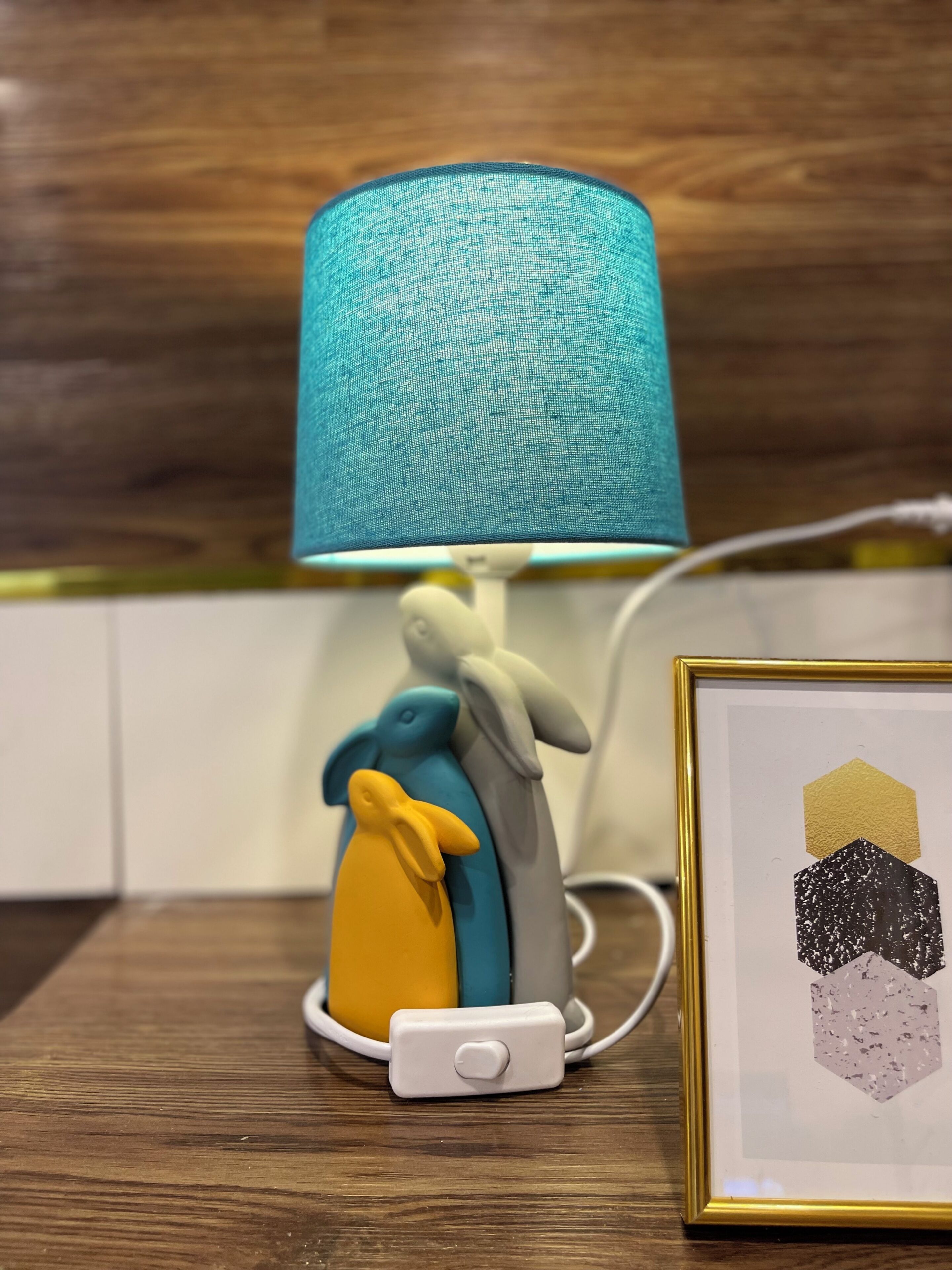 A teal lamp with a white base and a switch, placed on a wooden surface next to a framed artwork.