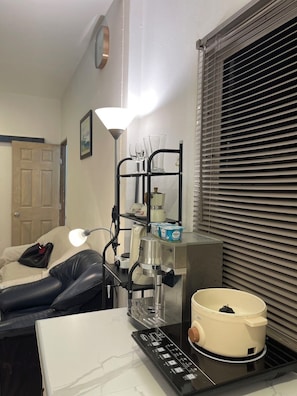 A hotel room with a bed, a coffee maker, a lamp, a shelf with glasses, and a window with blinds.