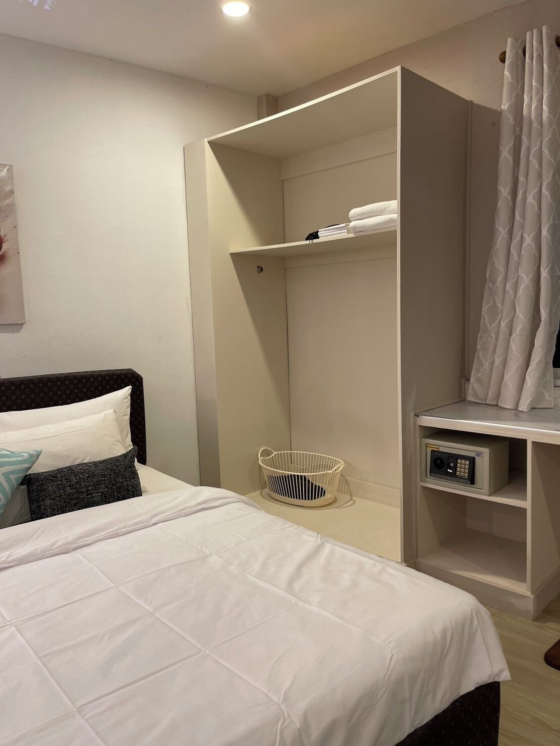 A hotel room with a bed, a white bedside table, a small white basket, a white shelf with towels, and a shower area with a white curtain.