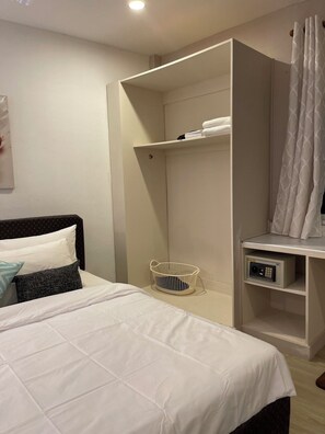 A hotel room with a bed, a white bedside table, a small white basket, a white shelf with towels, and a shower area with a white curtain. - Rural Stay Baan Mea Pen  Nature Retreat  (Chiang Rai)