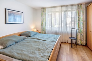 Ein Zimmer mit zwei Betten, einem gerahmten Bild an der Wand, einer Lampe auf einem Nachttisch und einem Sessel.