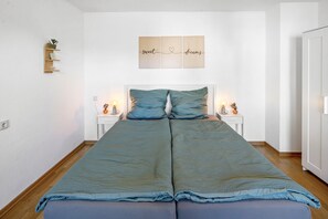 Cama doble con colcha y almohadas azules, dos mesitas de noche con lámparas y un estante en la pared con una planta.