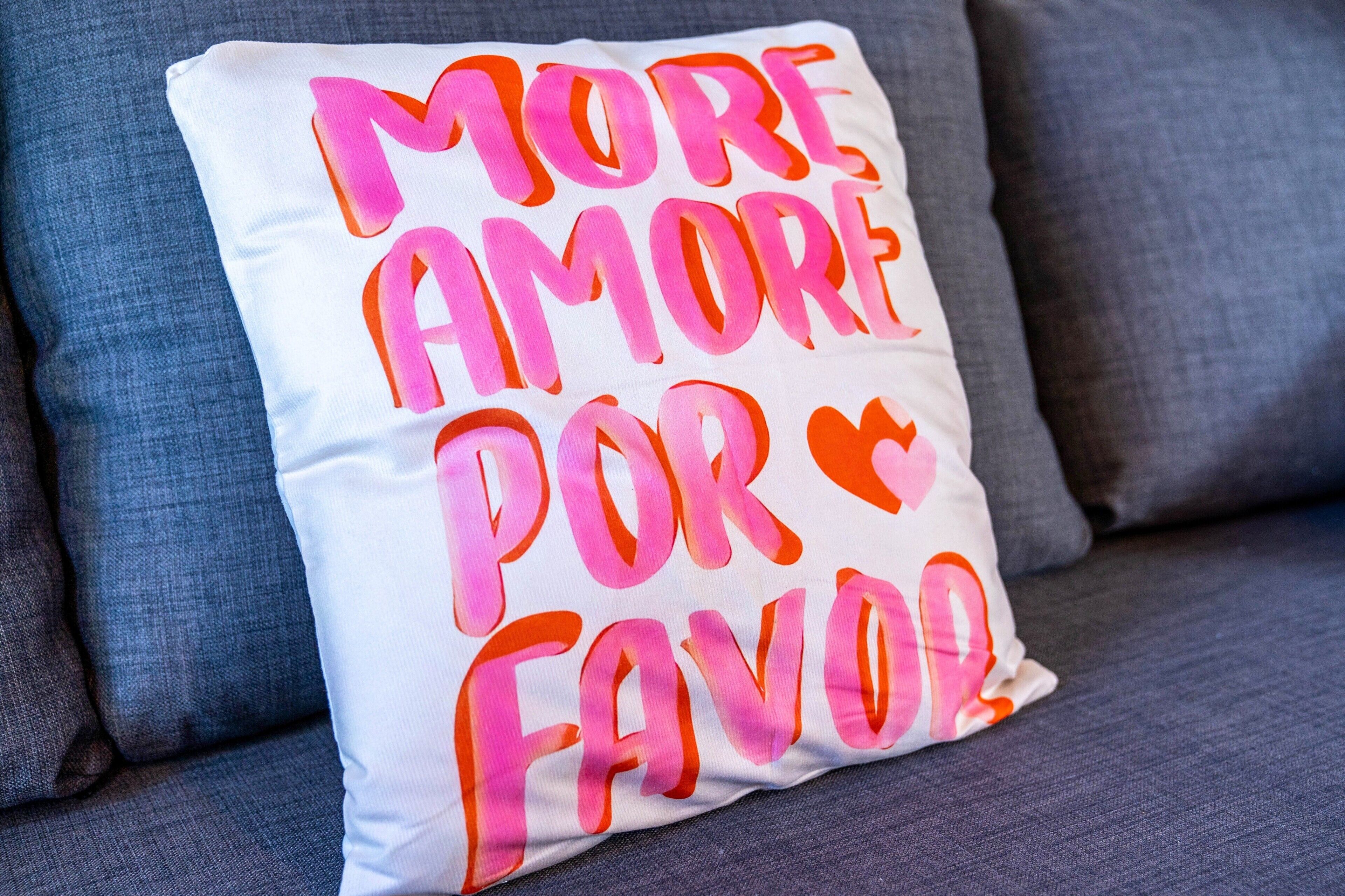 A pillow with the text 'MORE AMORE POR FAVOR' on a gray couch.
