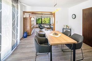Un interior moderno con una mesa de comedor de madera, sillas tapizadas en gris, una mesa de centro de vidrio y vistas a zonas verdes a través de amplios ventanales.