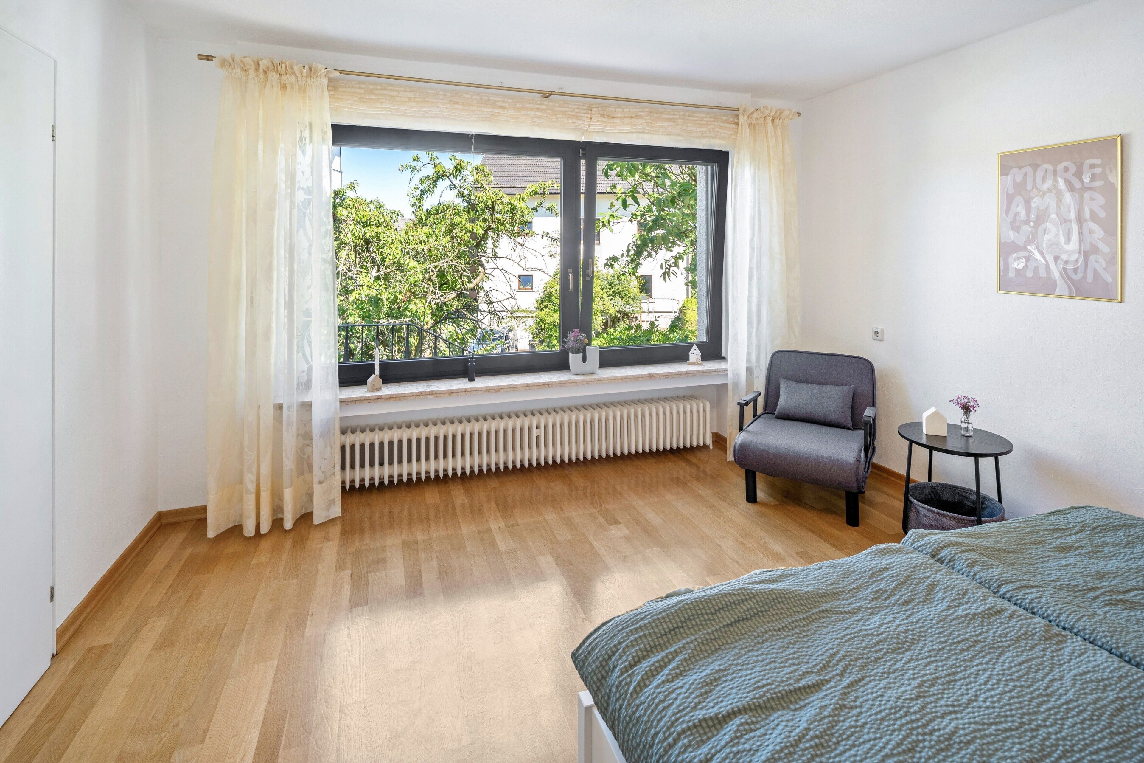 Ein Schlafzimmer mit großem Fenster, Heizkörper, Stuhl, Nachttisch und Bett.