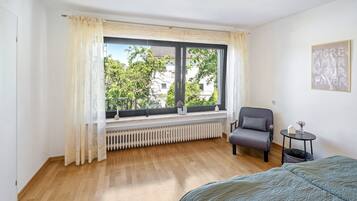 Ein Schlafzimmer mit großem Fenster, Heizkörper, Stuhl, Nachttisch und Bett.