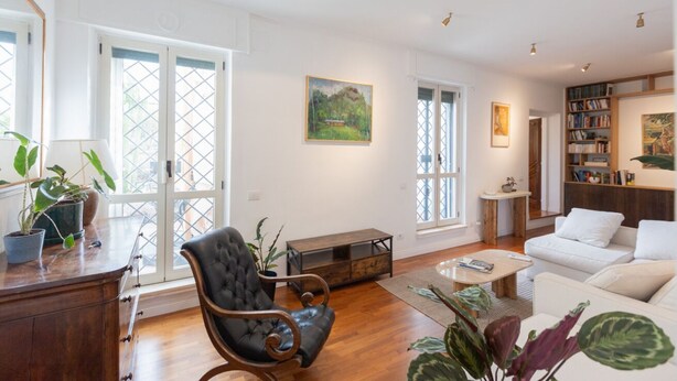 Ein geräumiges Wohnzimmer mit großem Fenster, einem Ledersessel, einem weißen Sofa, einem Couchtisch und einem Bücherregal.