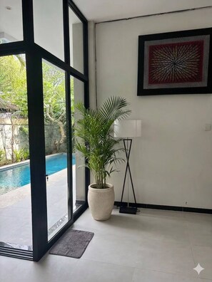 A room with a large glass door leading to a pool, a potted plant, and a framed artwork on the wall. - 3 BR, 250 Sqmt Villa, Terrace, 10 min Finns (Canggu)