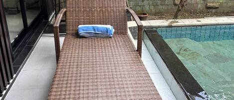 Un fauteuil de relaxation en osier, avec une serviette, placé près d’une piscine, avec un mur en pierre et de la verdure en arrière-plan.
