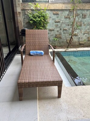 A wicker lounge chair with a towel, positioned near a pool with a stone wall and greenery in the background. - 3 BR, 250 Sqmt Villa, Terrace, 10 min Finns (Canggu)