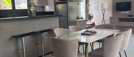 A modern kitchen with a built-in refrigerator, a dining area with a marble table and upholstered chairs, and a flat-screen TV mounted on the wall.