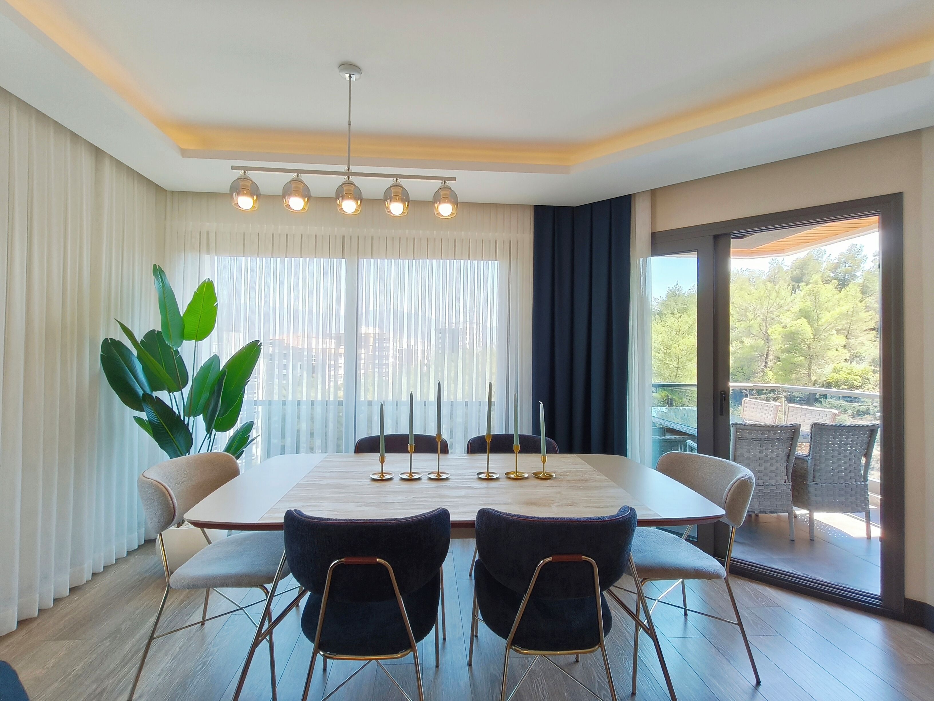 Un espace repas comprenant une grande table, six chaises, un lustre et une vue dégagée sur l’extérieur grâce à une grande fenêtre.