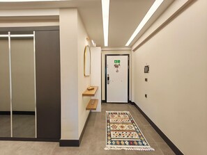 A hallway with a door marked for a lift call, a small shelf with a decorative item, and a patterned rug on the floor.