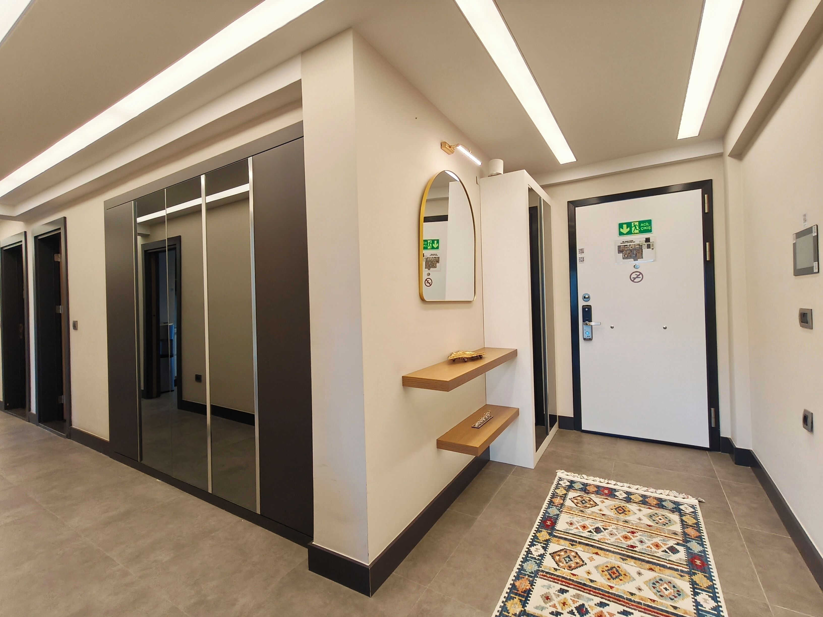 A hallway with a mirrored wall, a door with an emergency exit sign, and a patterned rug on the floor.