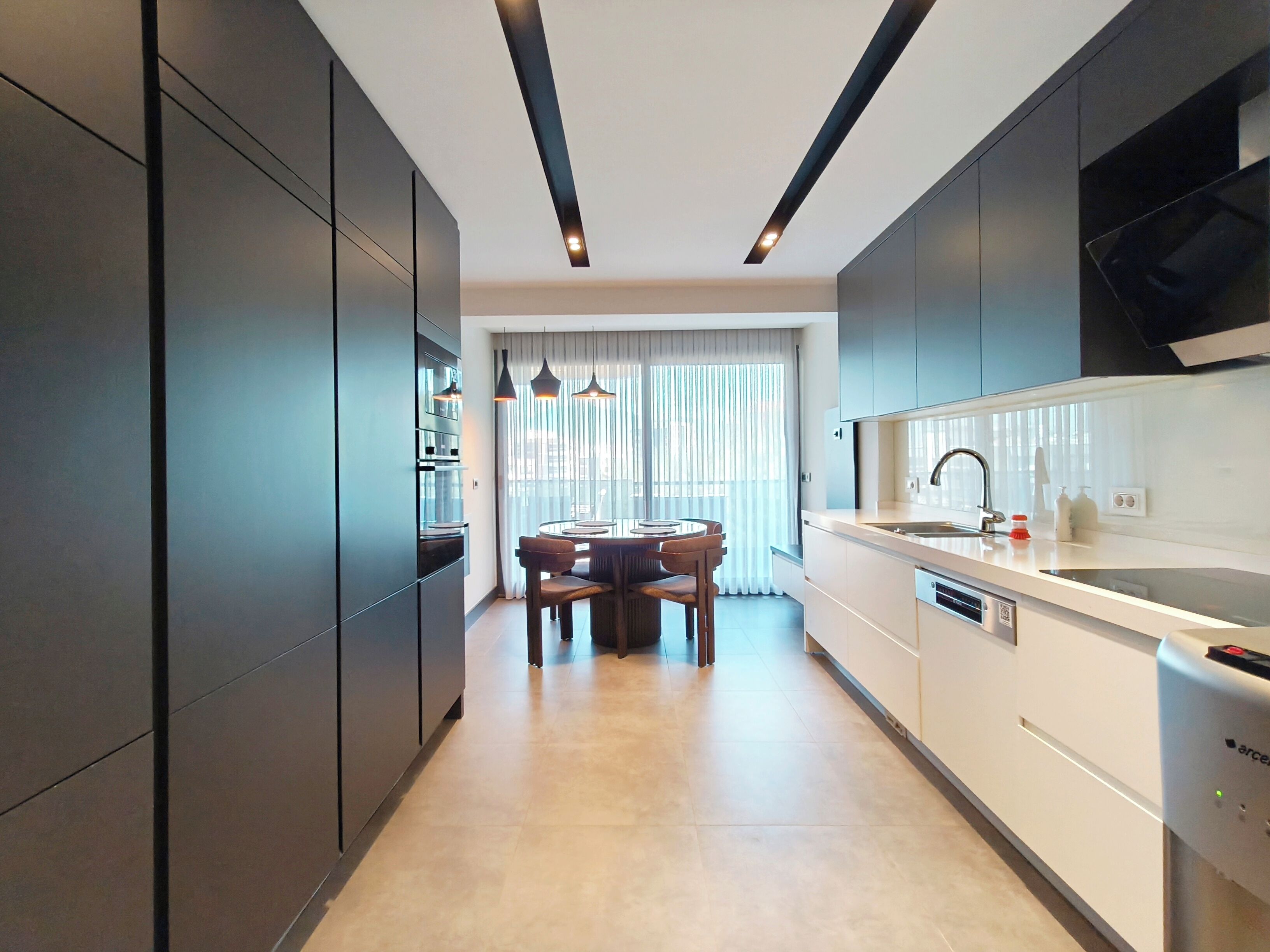 A modern kitchen with dark cabinetry, a dining area with a table and chairs, and a large window with sheer curtains.