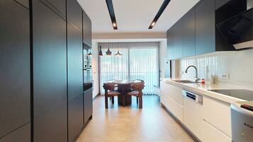 A modern kitchen with dark cabinetry, a dining area with a table and chairs, and a large window with sheer curtains.