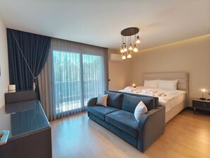 A modern hotel room with a large bed, a sofa, a desk, and a large window with curtains.