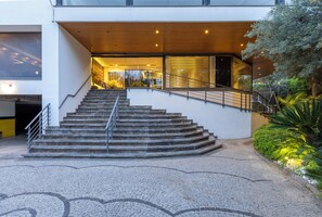 Un escalier menant à l’entrée d’un immeuble à l’architecture moderne, une zone pavée ornée d’un motif décoratif et un jardin bien entretenu où poussent différentes espèces de plantes.