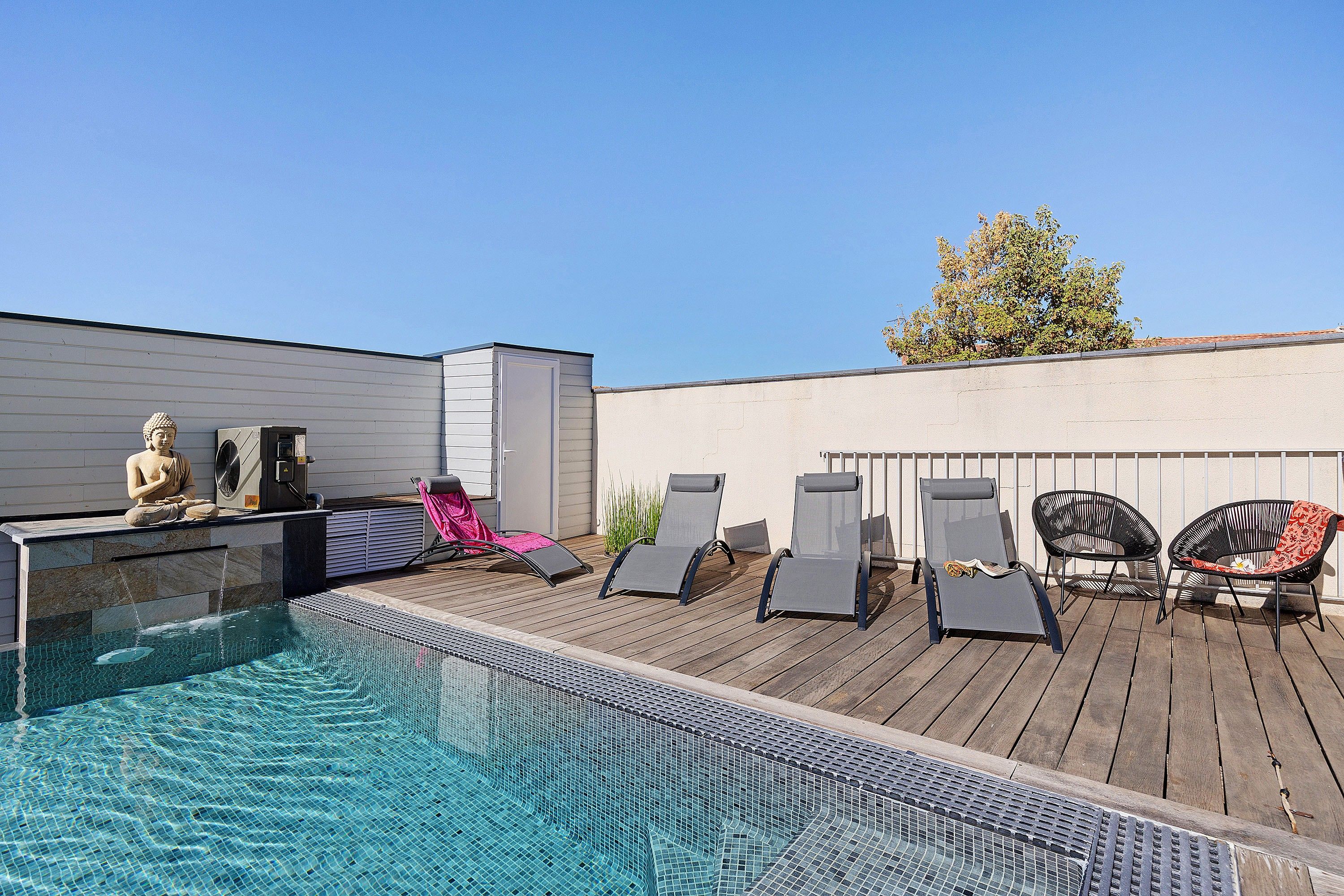 Uma área de piscina no terraço com uma estátua de Buda, espreguiçadeiras e céu azul límpido.