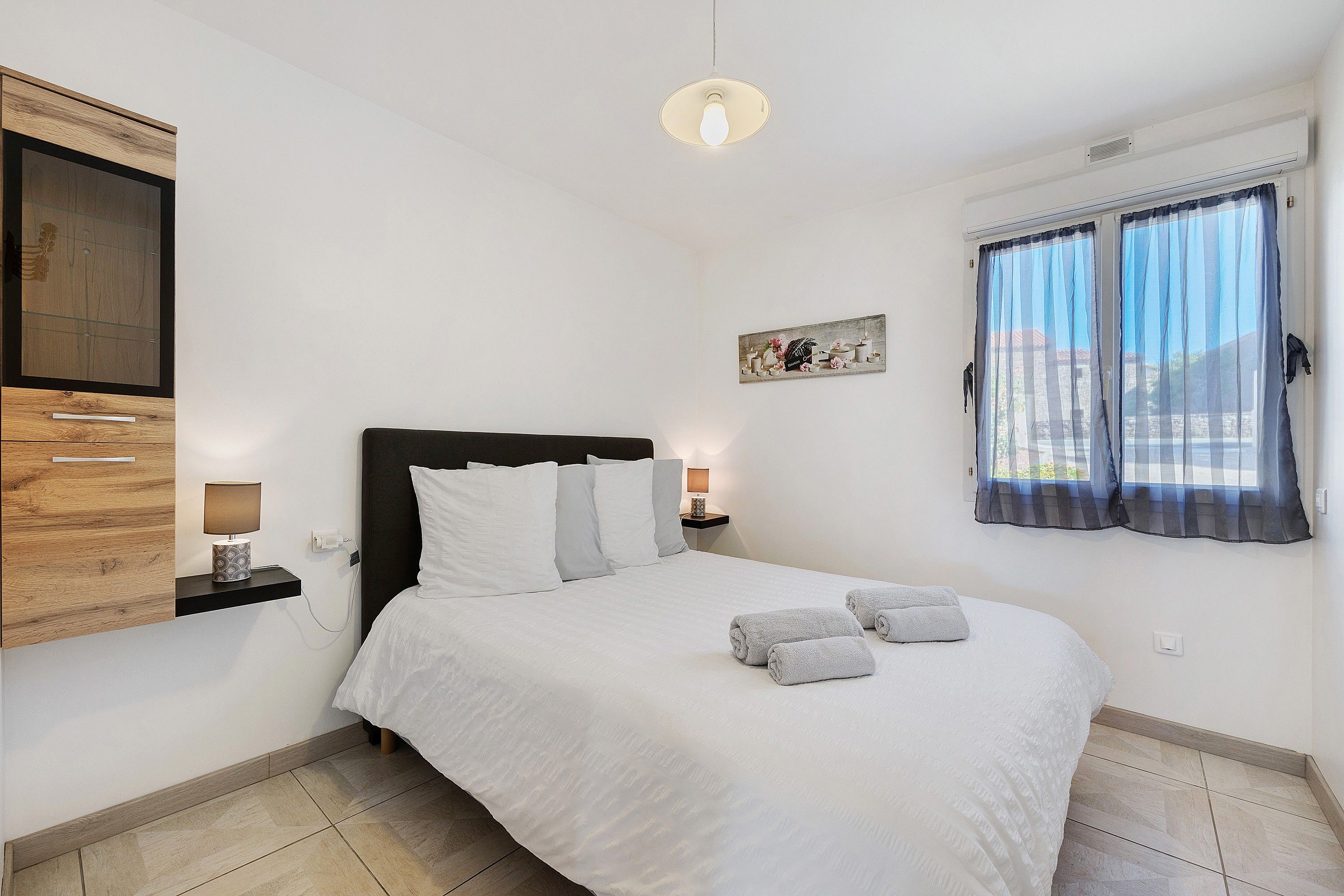 Un dormitorio con cama, mesitas de noche, un televisor montado en la pared, una ventana con cortinas y un cuadro.