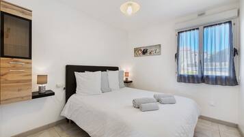 Un dormitorio con cama, mesitas de noche, un televisor montado en la pared, una ventana con cortinas y un cuadro.