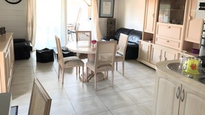 Una stanza ben illuminata con un tavolo da pranzo, sedie, un divano nero, un angolo cottura con pensili e una sveglia appesa al muro.