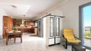 A modern kitchen with wooden cabinets, a dining area with a table and chairs, and a yellow armchair by a large window. - Solena - Garden Escape with Views near Chania (Tsikalaria)